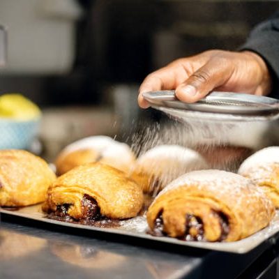 Close of a baker sprinkling powdered white sugar over croissants filled with chocolate in a baking tray